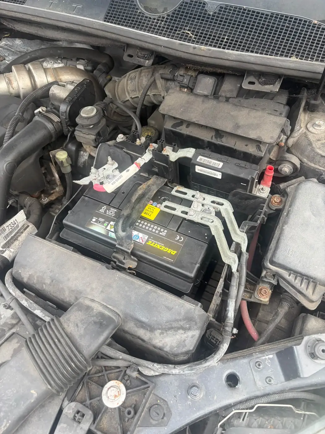 The old black car battery in the engine bay before the swap