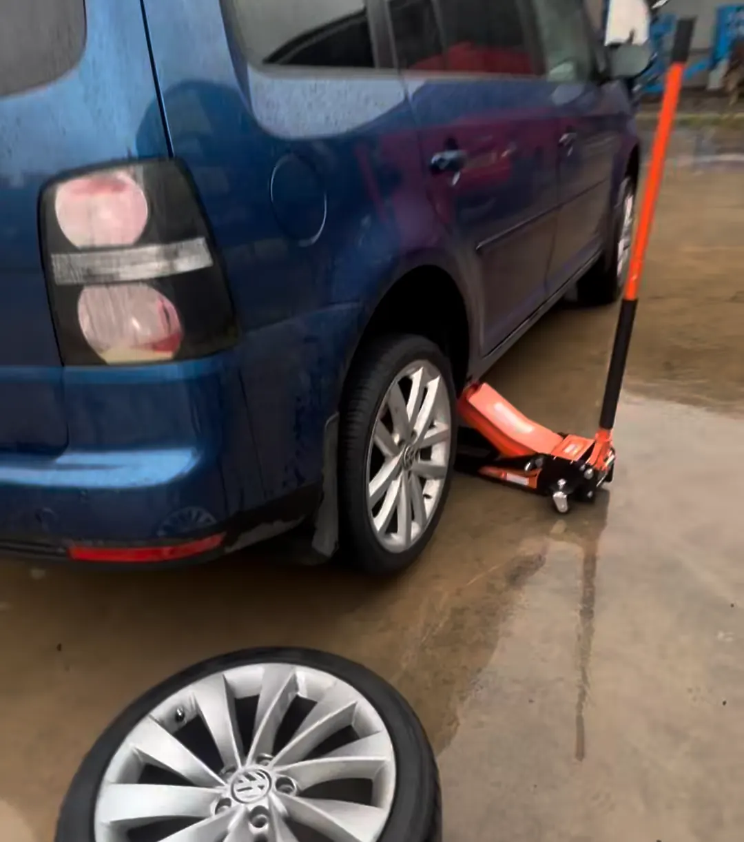 Blue VW with the rear wheel off, raised on an orange trolley jack in a wet yard, the alloy wheel lying in front